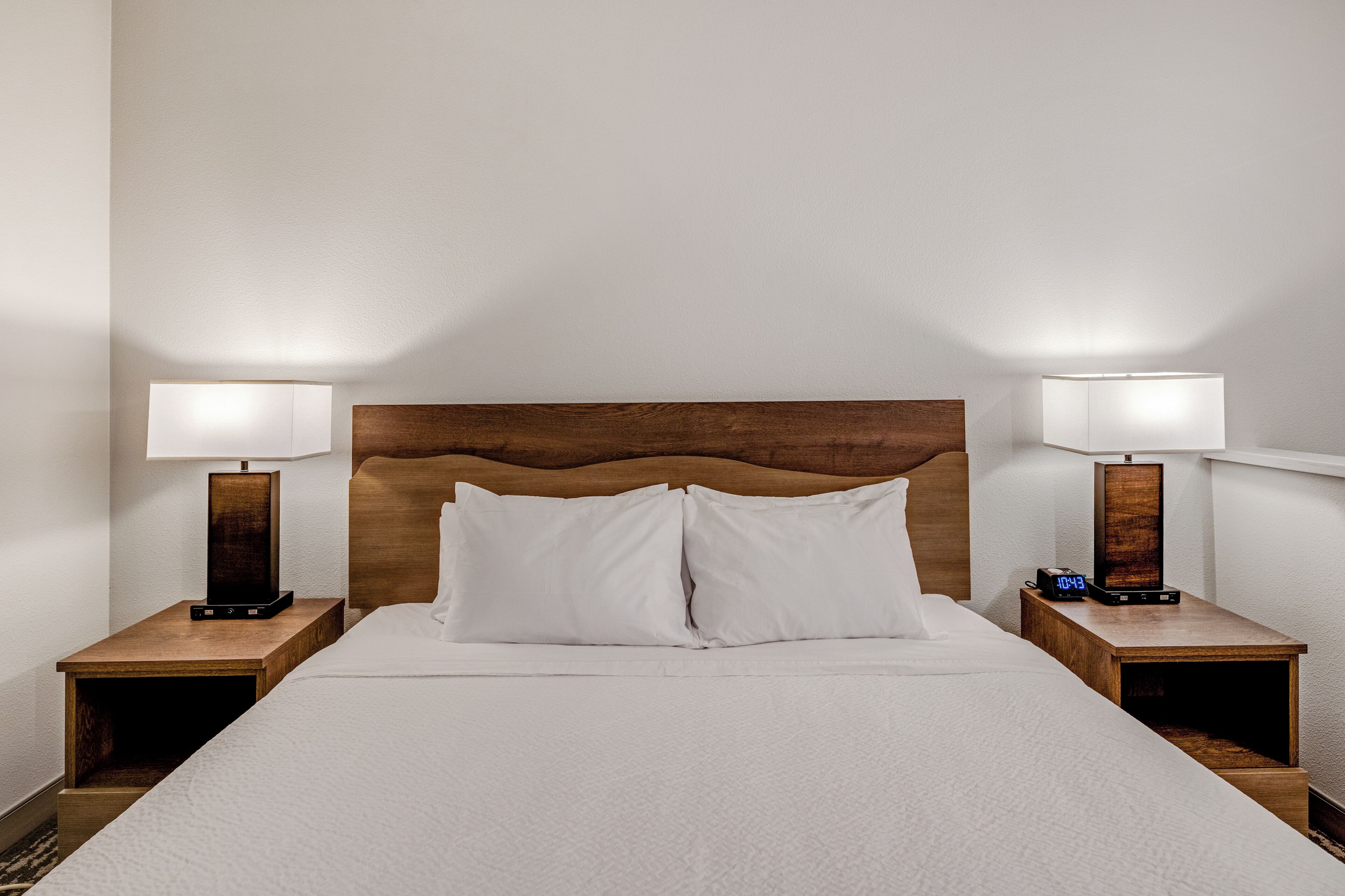 Quarto de hotel com cabeceira de madeira, duas mesas de cabeceira com abajures, cama com roupa de cama branca e um relógio digital em uma das mesas.