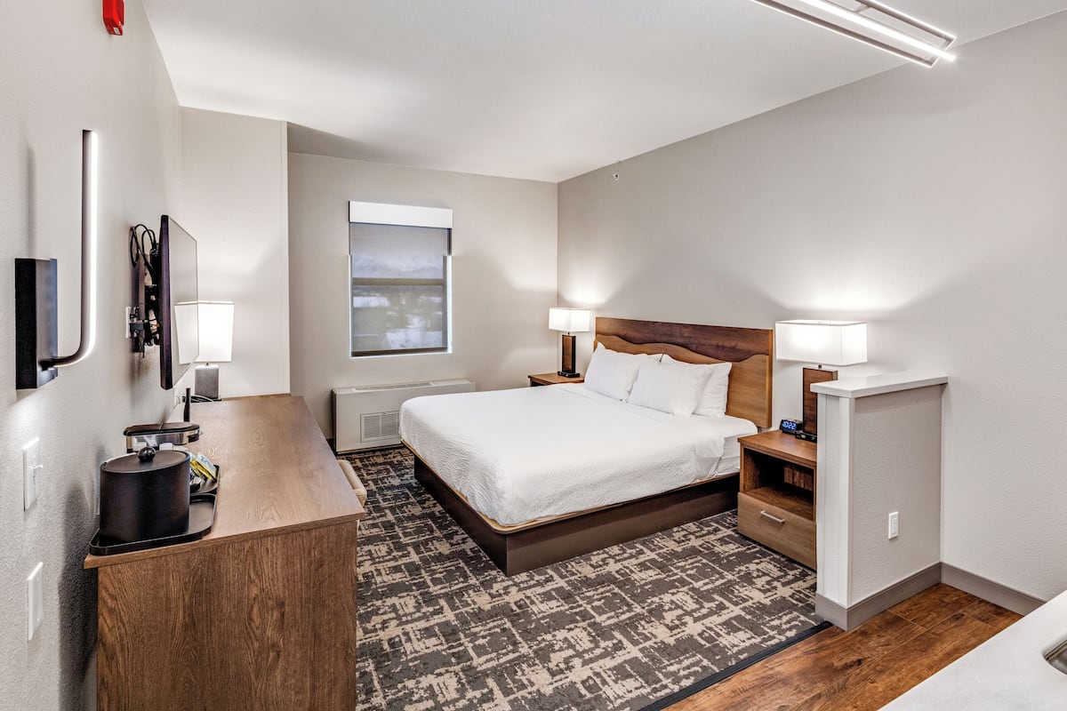 Ein modernes Hotelzimmer mit Bett, Nachttischen, Schreibtisch, Spiegel, Fenster und einer Deckenleuchte.