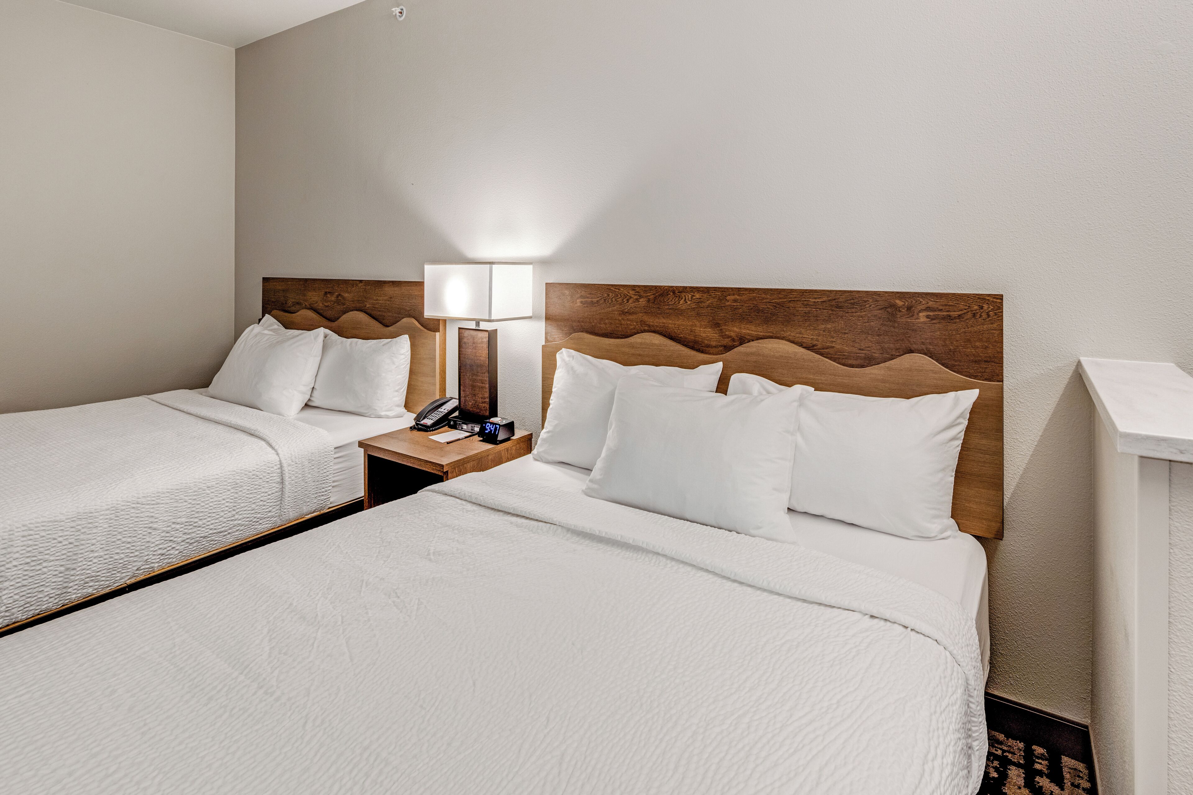 A hotel room with two beds, a nightstand with a lamp, a phone, and a clock.