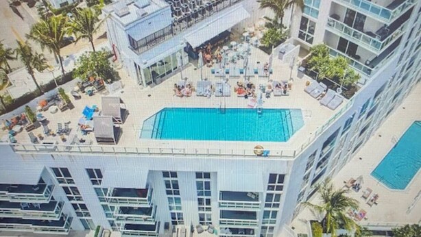 An aerial view of a hotel with a rooftop pool surrounded by lounge chairs and umbrellas, adjacent to a high-rise building with balconies.