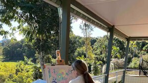 Une personne est en train de peindre sur un chevalet, sur une terrasse en bois, avec vue sur une piscine et sur la verdure environnante.