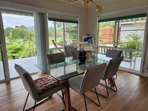 A dining area with a glass table and chairs, large windows with a view of greenery, and a ceiling light fixture.