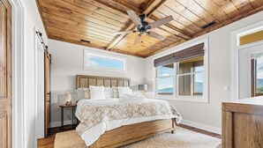 A bedroom with a wooden ceiling, a bed with a white and beige bedspread, a nightstand with a lamp, and a window with a view of the outdoors.