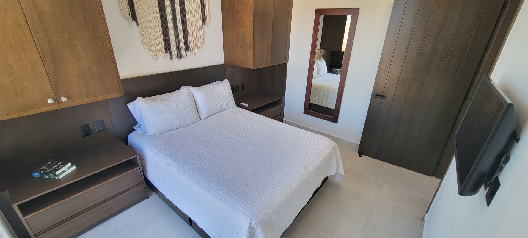 Quarto com cama, guarda-roupa de madeira, mesas de cabeceira, espelho e televisão.