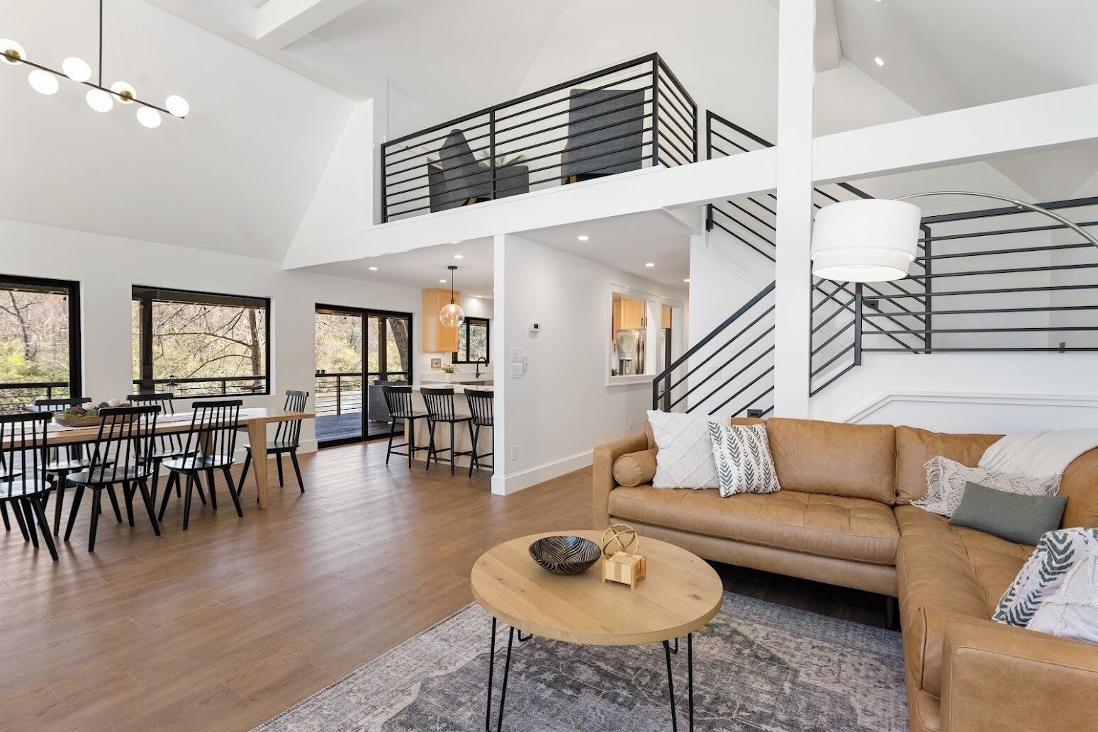 A spacious living area with a high ceiling, a dining area with a large window, and a staircase leading to an upper level.