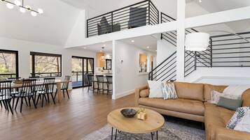 A spacious living area with a high ceiling, a dining area with a large window, and a staircase leading to an upper level.
