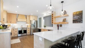 Eine moderne Küche mit Holzschränken, Edelstahlgeräten, einer großen Kücheninsel mit Barhocker, Pendelleuchten und einem Bild an der Wand.