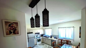 Eine Küche mit weißen Schränken, einem Kühlschrank, einem Backofen, einer Spüle und einem Esstisch mit Stühlen. Es gibt Pendelleuchten und ein Fenster mit Vorhängen.