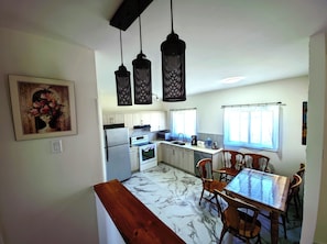 A kitchen with white cabinets, a refrigerator, an oven, a sink, and a dining table with chairs. There are hanging lights and a window with curtains.