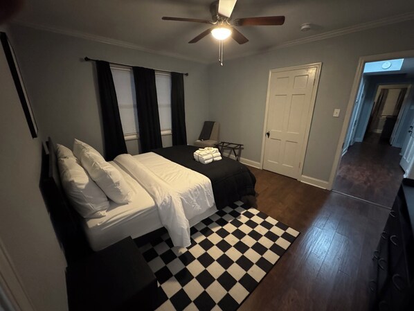Un dormitorio con cama, alfombra a cuadros blanco y negro, ventilador de techo y puerta que da a otra habitación.
