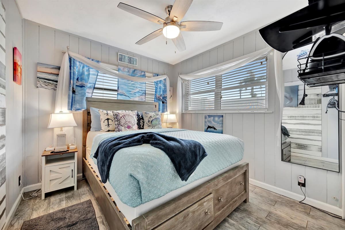 A bedroom with a bed, a nightstand, a ceiling fan, a window with blinds, and a wall-mounted mirror.