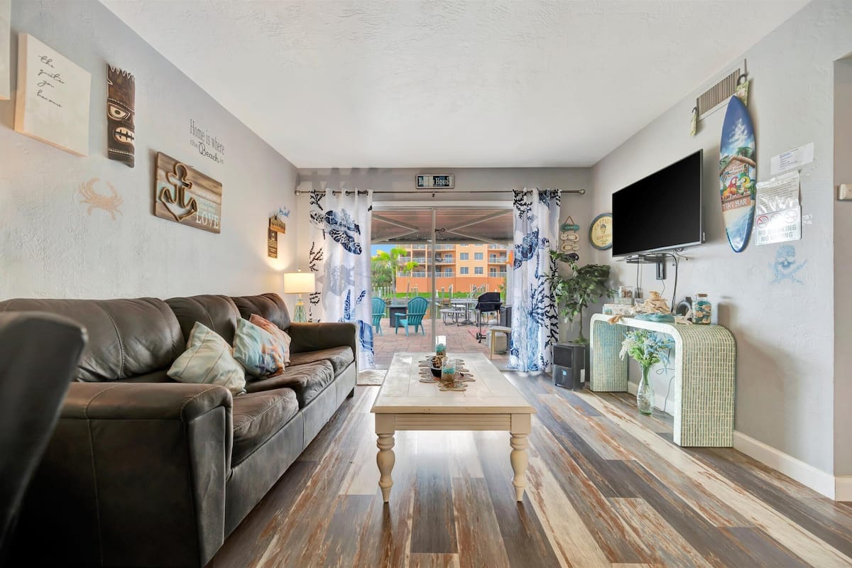 A living room with a leather sofa, a coffee table, and a TV. There are decorative items on the walls and a view of the outside.