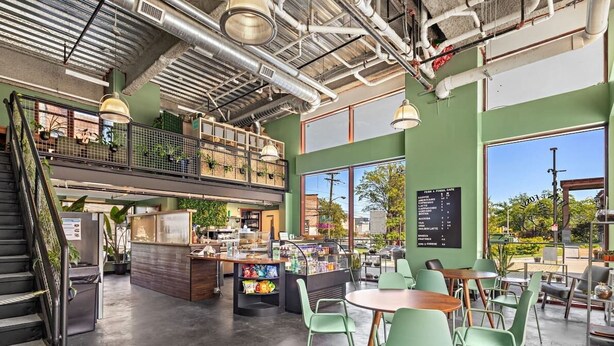 Ein geräumiges Café mit hoher Decke, freiliegenden Lüftungsrohren und einer Galerie. Es gibt Tische und Stühle, eine Theke mit Speisekarte und eine Treppe, die zum oberen Bereich führt.