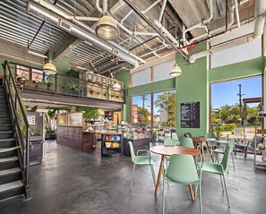 Un café spacieux avec un plafond haut, des conduits apparents et une mezzanine. On y trouve des tables et des chaises, un comptoir avec un menu et un escalier menant à l’étage supérieur.