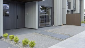 A modern building with a grey facade, a large glass door entrance, and a small garden area with shrubs.