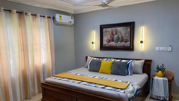 A bedroom with a wooden bed, a white rug, a ceiling fan, and a painting on the wall. - 3-bedroom house at East Legon with 24hr security and solar powered lights (Accra)