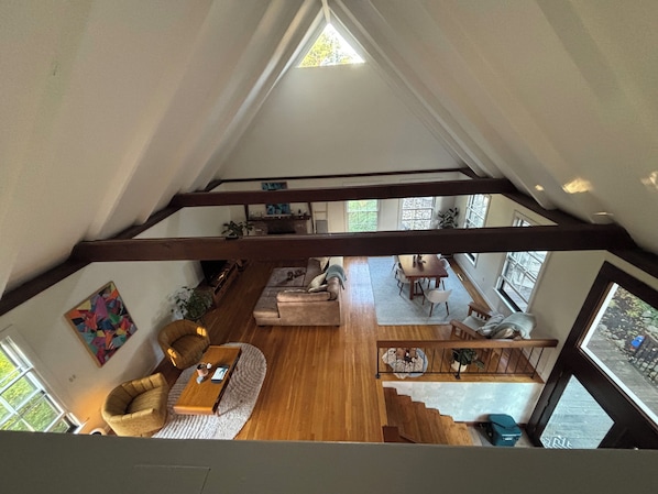 Ein geräumiges Loft mit hohen Decken, freiliegenden Holzbalken, einer gemütlichen Sitzecke mit Sofa und Sesseln, einem Esstisch mit Stühlen und großen Fenstern mit Blick ins Freie.