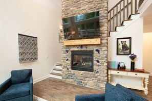 A living room with a stone fireplace, a staircase, and a mounted television.
