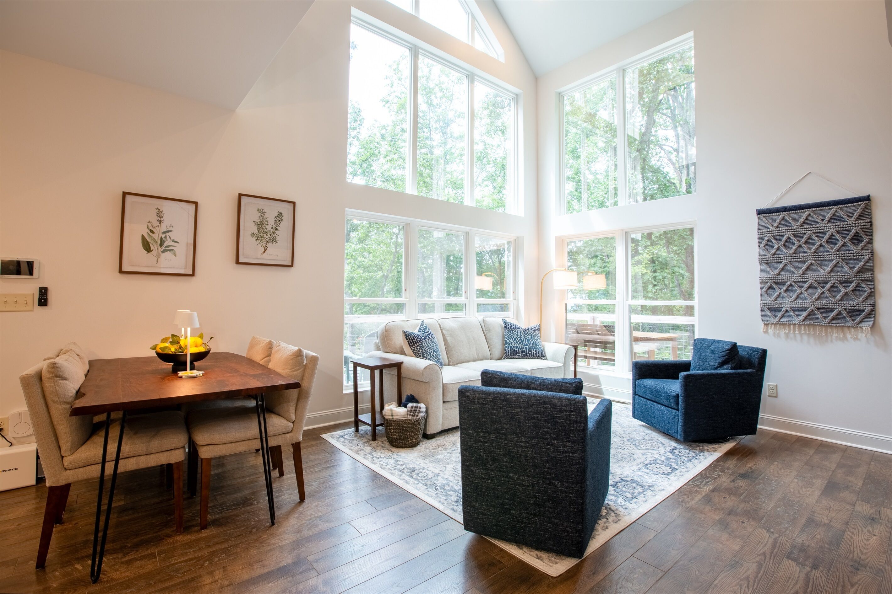Un salon spacieux avec une table à manger en bois, un canapé et un fauteuil bleu. De grandes fenêtres laissent entrer la lumière naturelle et offrent une vue sur l’extérieur.