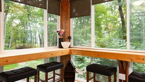 A corner seating area with a wooden bench and a table, large windows with a view of a forest.