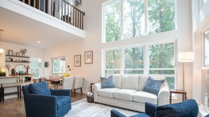 A spacious living room with a high ceiling, large windows, a white sofa, and blue armchairs.