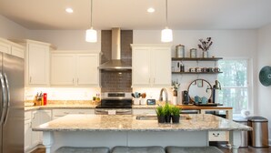 A modern kitchen with white cabinets, a stainless steel refrigerator, a central island with bar stools, pendant lighting, and a window providing natural light.