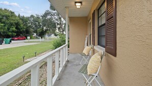 A covered porch with two chairs and a bench, a street view, and a red car parked on the side. - Fully Renovated & Private 2-bedroom home in Zephyrhills with Porch (Zephyrhills)