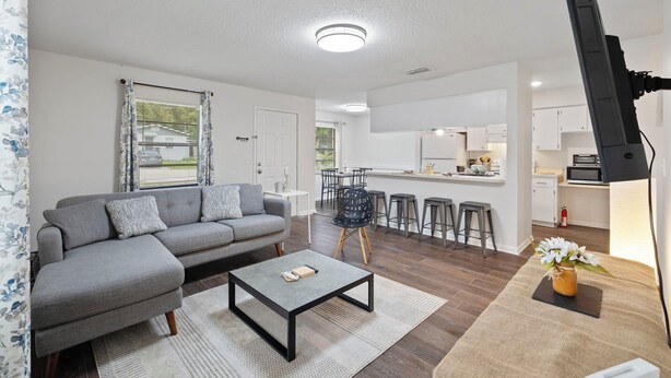 A modern living room with a grey sofa, a coffee table, and a kitchen area in the background. - Fully Renovated & Private 2-bedroom home in Zephyrhills with Porch (Zephyrhills)