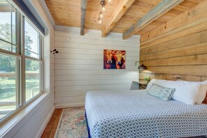 Une chambre avec un plafond en bois, un lit recouvert d’une literie à motifs, un tableau représentant des chevaux et une fenêtre donnant sur un paysage verdoyant.
