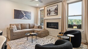A living room with a sectional sofa, a fireplace, a ceiling fan, and a large window with a view of trees.