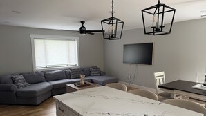 Ein modernes Wohnzimmer mit grauem Sofa, einem Esstisch und einer Küchenzeile mit weißen Schränken und einer Marmorarbeitsplatte.