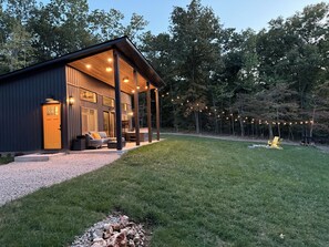 Una cabaña moderna con un porche cubierto, mobiliario exterior y luces colgantes.