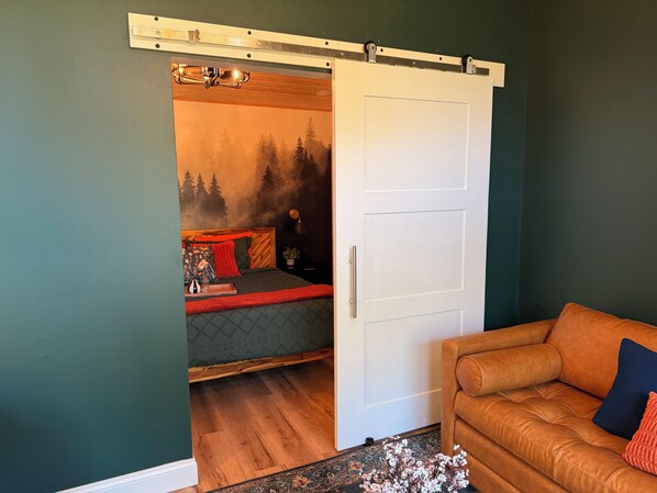 Una habitación con puerta corrediza que da a un dormitorio con cama, mesita de noche y un mural en la pared.