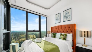 A modern hotel room with a large bed, a view of the city, and two framed artworks on the wall.