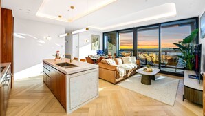 A modern living space with a kitchen, dining area, and a large window offering a view of the sunset.