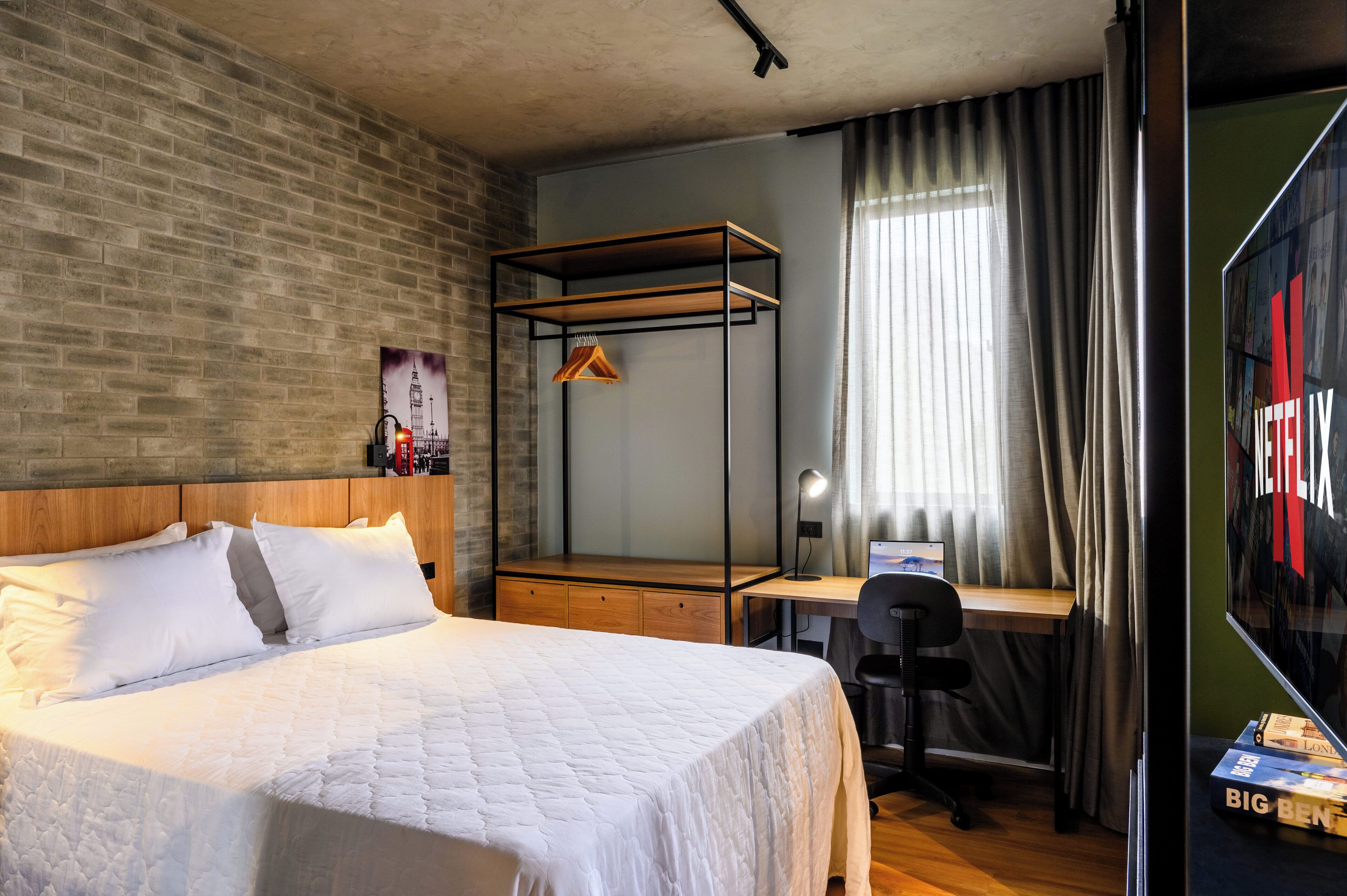 A modern hotel room with a brick wall, a wooden bed, a desk with a chair, and a large window with curtains.