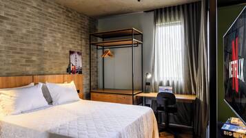 A modern hotel room with a brick wall, a wooden bed, a desk with a chair, and a large window with curtains.