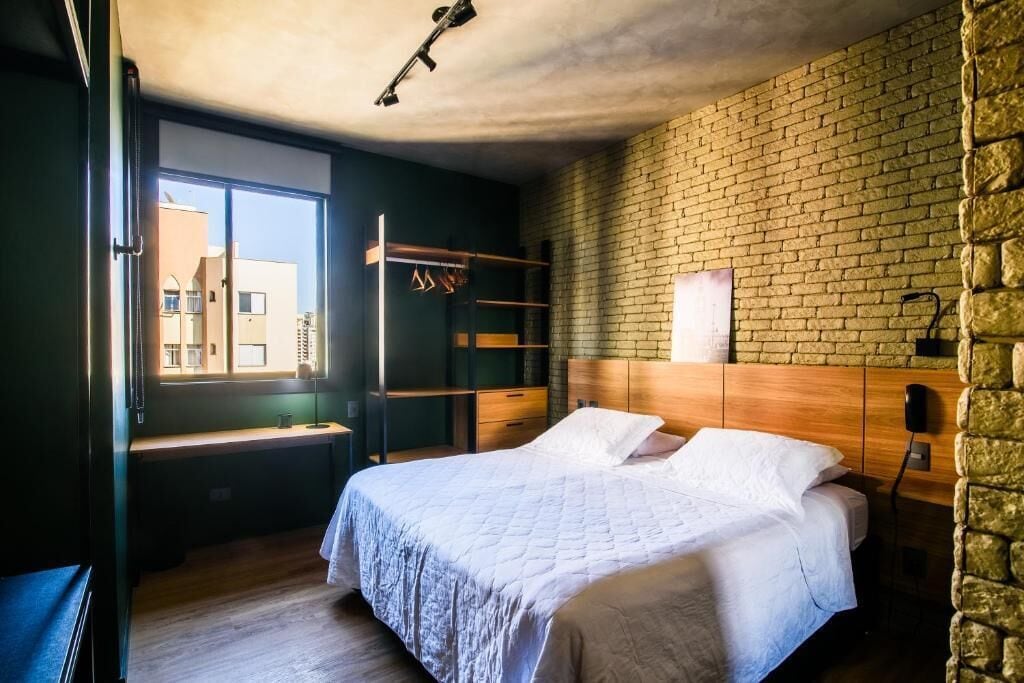 A bedroom with a brick wall, a bed with white bedding, a wooden headboard, and a window with a view of buildings.