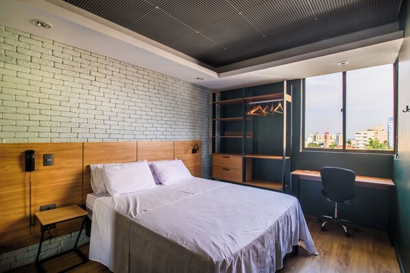 A modern hotel room with a brick wall, a wooden headboard, a bed with white linens, a desk with a chair, and a large window with a city view.