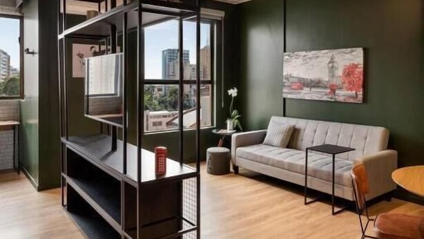 A modern interior with a high ceiling, a black metal shelving unit, a grey sofa, and a cityscape view through large windows. - LUIX LONDRINA FLAT (Londrina)