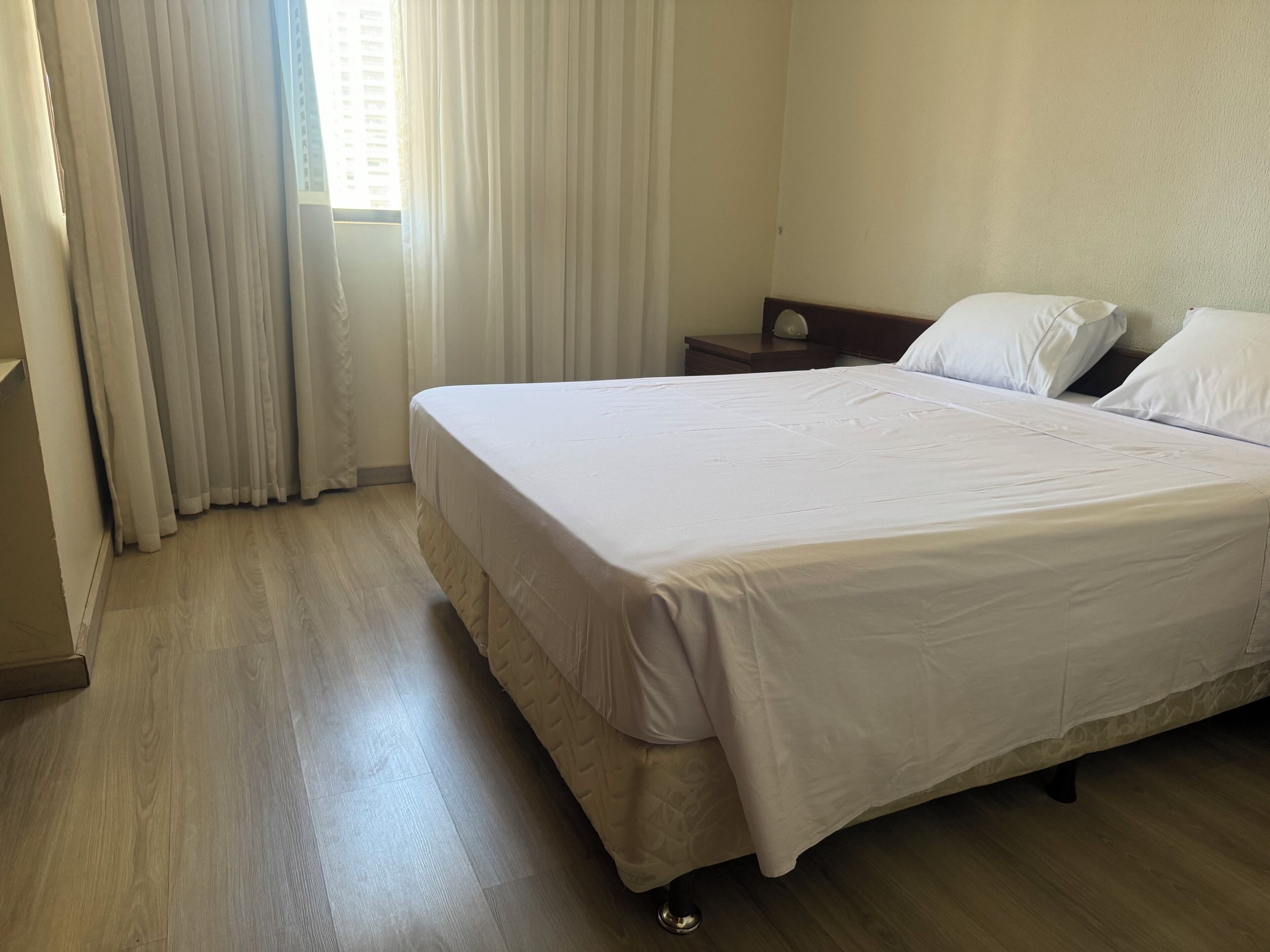 A neatly made bed with a white bedspread and pillows, a wooden floor, a nightstand with a lamp, and a window with curtains.