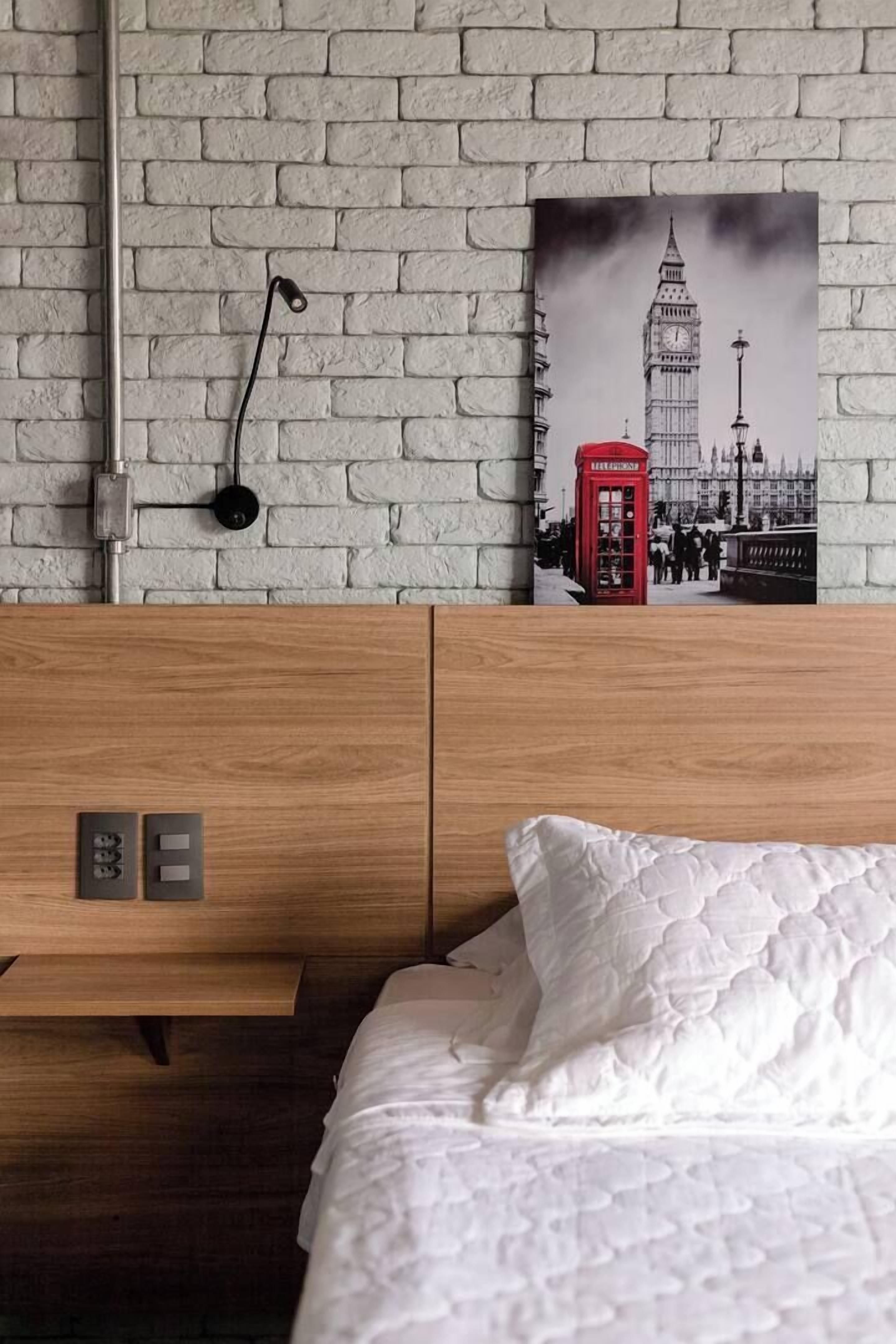 A hotel room with a brick-patterned wall, a bed with white bedding, a wooden headboard, a red telephone booth artwork, and a wall-mounted light fixture.