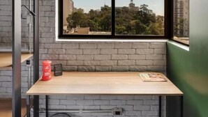A small, modern room with a brick wall, a large window with a black frame, a wooden desk, and a black trash bin. - LUIX LONDRINA FLAT (Londrina)