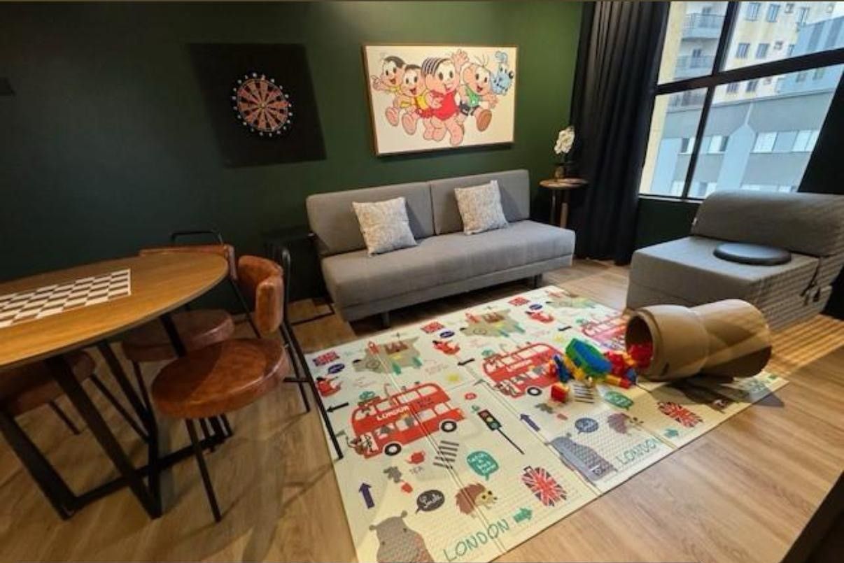 Quarto com uma mesa de jantar, sofá, tapete infantil e janela com vista para edifícios.