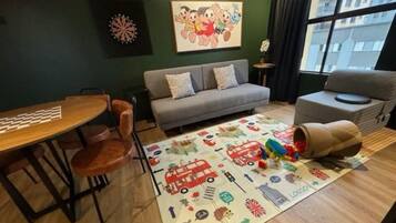 Quarto com uma mesa de jantar, sofá, tapete infantil e janela com vista para edifícios.