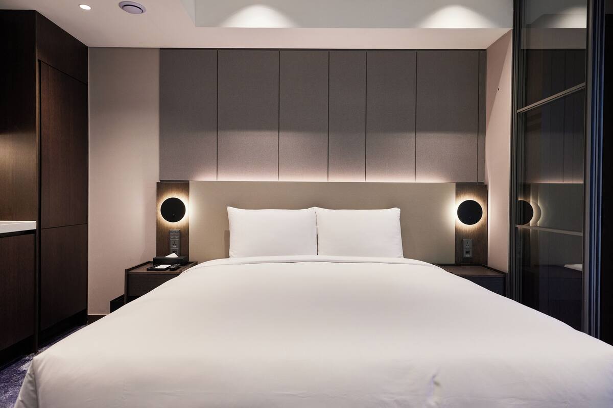 Habitació d'hotel moderna amb un llit gran, panell de llit integrat, làmpades laterals i una paret divisòria de vidre.