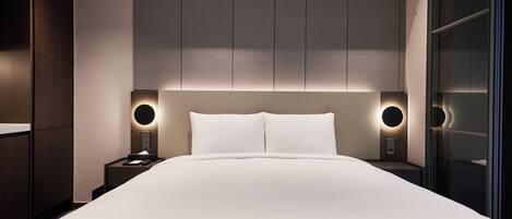 A modern hotel room with a large bed, built-in headboard, bedside lamps, and a glass partition wall.