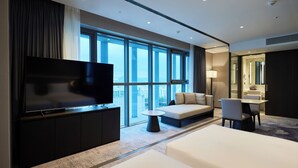A modern hotel room with a large flat-screen TV, a sofa, a dining table, and a large window offering a city view. - HOTEL HLB (Busan)