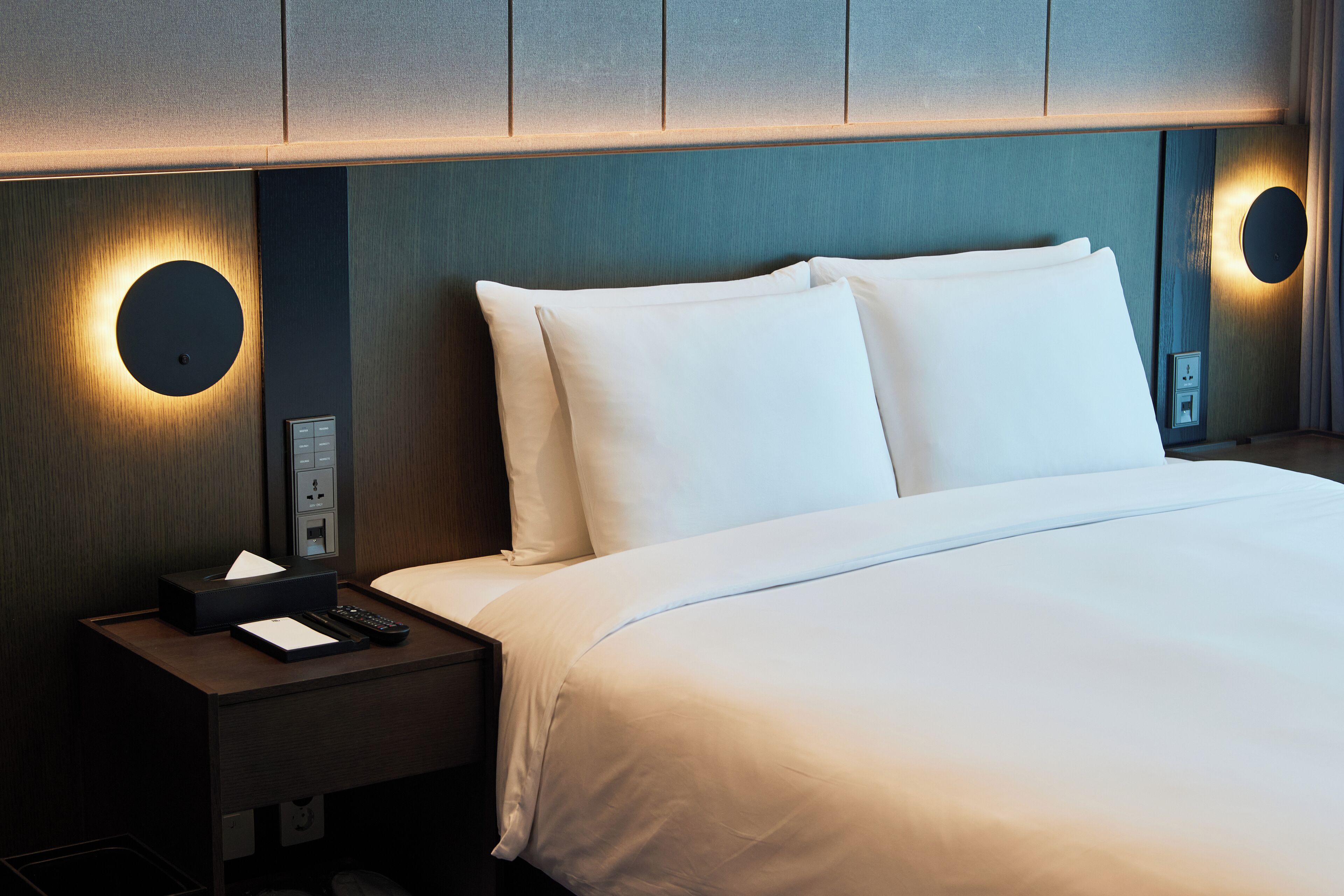 Ein Hotelzimmer mit einem ordentlich bezogenen Bett, einem Nachttisch mit Fernbedienung und einer an der Wand befestigten Lampe.