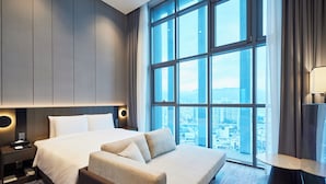 A modern hotel room with a large bed, a sofa, a nightstand, and a view of the city through large windows. - HOTEL HLB (Busan)
